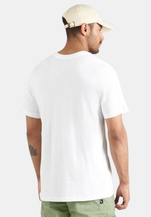 Hombre con camiseta blanca lisa, gorra beige y pantalones verdes, de perfil hacia la izquierda con la espalda mayormente visible contra un fondo claro.