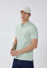 Light green polo shirt with short sleeves and a collar, featuring two buttons. Worn with a beige cap, standing against a white brick wall.