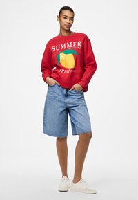 Sudadera roja con gráfico de "SUMMER MANGO TREE FRUIT JUICE", combinada con pantalones cortos de mezclilla desgastados y zapatillas blancas. Atuendo casual.
