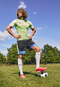 Cămașă sportivă verde și albă cu un model, pantaloni scurți negri cu accente albastre, șosete albe și încălțări de fotbal roșii pe iarbă, cu o minge de fotbal în apropiere.