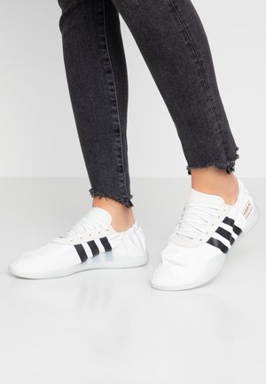 Personne portant des baskets blanches avec des rayures noires et un jean skinny noir à ourlet effiloché, debout sur un sol blanc lisse.