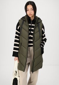 Gilet vert olive rembourré avec capuche, doté d'une fermeture zip, d'un design matelassé et de bretelles noires, porté sur un haut à rayures noir et blanc.