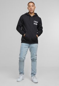 Sort zip-up hoodie med frontlommer, der har teksten "Escape from Reality" i hvid. Sammen med lyseblå jeans og hvide sneakers.