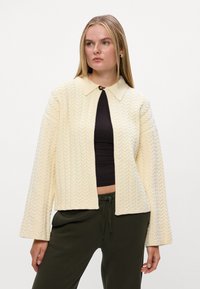 Kremowy sweter dzianinowy o prążkowanej strukturze, z kołnierzem i zapięciem na guziki. Posiada szerokie rękawy i krótki krój.