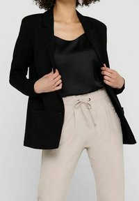 Černý blazer se strukturovanými klopami a dvěma předními kapsami, v kombinaci s černým saténovým topem a béžovými tepláky s elastickým pasem.