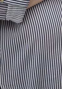 Nærbillede af en person iført en blå og hvid vertikalt stribet skjorte med opkrøllede ærmer og en guldbutton.