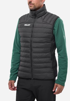 Homme portant un gilet sans manches matelassé noir avec fermeture éclair sur un haut vert à manches longues, debout devant un fond blanc.