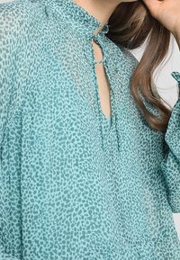 Tyrkysová blůza z lehkého, průhledného materiálu s tečkovaným vzorem. Má nabíraný výstřih a malý klíčový otvor vepředu.