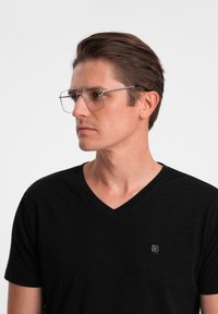 Przezroczyste prostokątne okulary z cienką srebrną oprawą, noszone przez osobę w czarnej koszulce z dekoltem w serek, prezentujące minimalistyczny design.