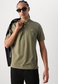 Tricou polo verde măsliniu cu mâneci scurte și un guler cu trei nasturi. Prezintă un mic logo pe piept. Modelul ține o jachetă din piele neagră.