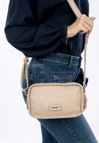 Borsa a tracolla in pelle beige con dettagli intrecciati, forma rettangolare e tracolla regolabile. Indossata sui fianchi con jeans in denim.