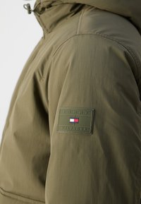 Manche de veste vert olive avec un patch logo Tommy Hilfiger de couleur assortie représentant un drapeau rouge, blanc et bleu.