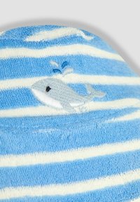 Világoskék és fehér csíkos kalap, puha törölköző anyagból, amelyen egy hímzett szürke bálna látható vízpárás részlettel.