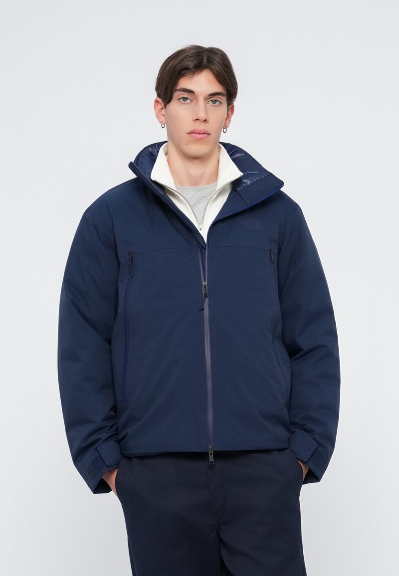 Veste isolante bleu marine avec un col haut, fermeture éclair à l'avant et deux poches avant. Présente une doublure intérieure blanche et des poignets texturés.