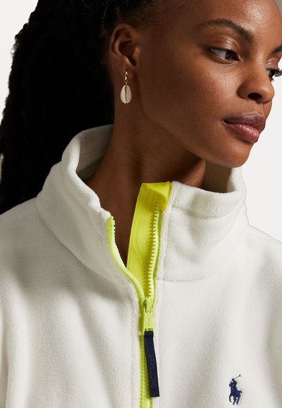 Weißer Fleece-Jacke mit Stehkragen, ausgestattet mit einem leuchtend gelben Reißverschluss und einem marineblauen Logo-Detail. Goldene muschelförmige Ohrringe werden getragen.