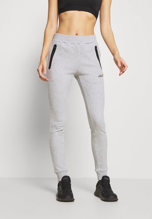 Kvinde iført lysegrå joggingbukser med sorte lynlåslommer, sort sports-bh og sorte sneakers, stående mod en ensfarvet hvid baggrund.