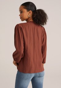 Blouse marron texturée avec des manches bouffantes trois-quarts, un col rond et une coupe légèrement ample, associée à un jean bleu.