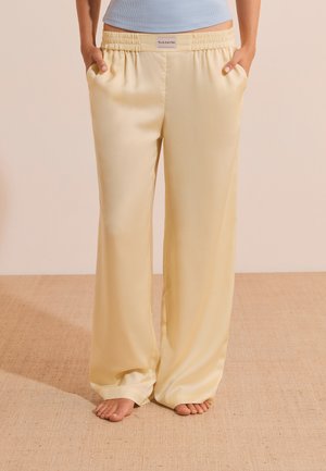 Personne portant un pantalon ample en satin jaune clair avec taille élastique et poches, debout pieds nus sur un sol beige texturé.