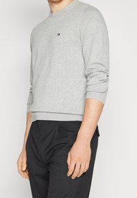 Uomo che indossa un maglione a maniche lunghe grigio chiaro con un piccolo logo sul petto e pantaloni plissettati neri, tagliati a metà faccia e a metà coscia.
