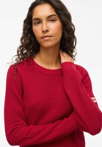 Pull en tricot de couleur rouge profond avec un motif texturé en nid d'abeille. Présente un col rond et des poignets côtelés. Manches longues, tissu doux.