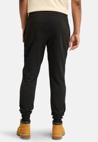 Schwarze Sweatpants mit elastischem Bund und Bündchen an den Knöcheln, von hinten gezeigt. Der Träger trägt beige Stiefel. Ledrige Stofftextur.