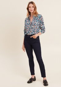 Camicia button-up blu con un motivo di foglie multicolore, abbinata a jeans slim-fit blu scuro e mocassini neri, su uno sfondo neutro.