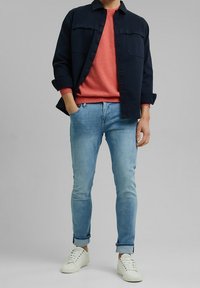 Marineblaues Button-up-Hemd über einem korallenfarbenen Crewneck-Pullover, kombiniert mit hellblauen Jeans und weißen Sneakers, zeigt einen lässigen, geschichteten Look.