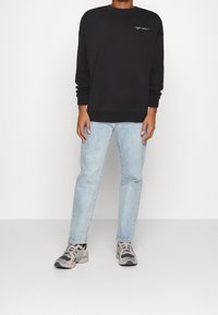 Sudadera negra con cuello redondo, puños acanalados y logo bordado en el pecho, combinada con vaqueros de mezclilla azul claro y zapatillas multicolores.