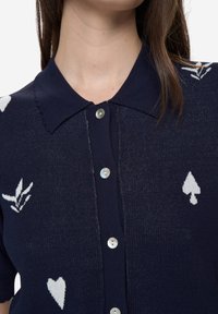 Tmavě modrý pletený kardigan s polokošilovým límcem, zdobený bílými vzory srdcí a pik. Perlové knoflíky dodávají detail na přední straně.