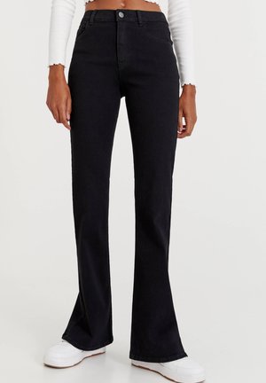 Smaltsiddende sorte jeans med let vidde, båret med hvide sneakers og en cropet hvid top med lange ærmer, der viser maven.