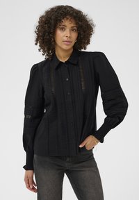 Femme aux cheveux bouclés portant une blouse noire à boutons avec des panneaux en dentelle et des manches bouffantes, associée à un jean gris foncé et un bracelet en or.