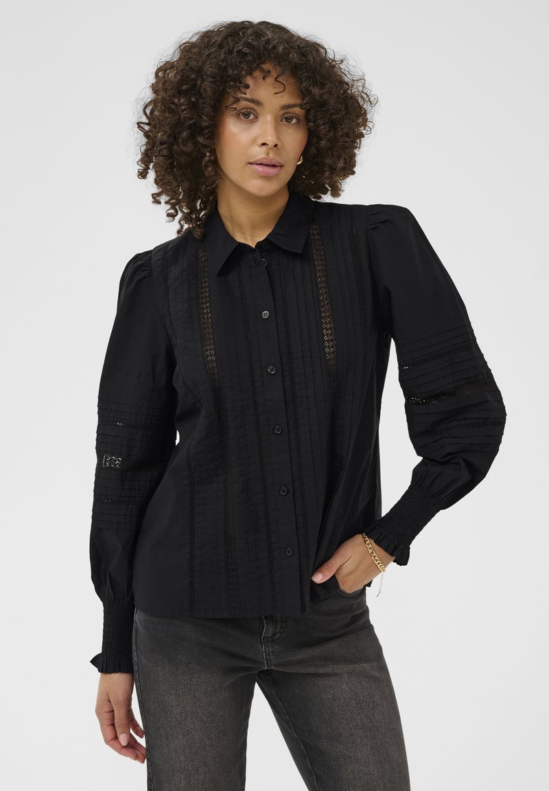 Femme aux cheveux bouclés portant une blouse noire à boutons avec des panneaux en dentelle et des manches bouffantes, associée à un jean gris foncé et un bracelet en or.