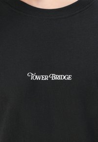 Tricou negru din bumbac cu guler la baza gâtului, având text brodat alb "TOWER BRIDGE" într-un font decorativ centrat pe piept.