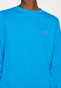 Sudadera azul de algodón con cuello redondo y mangas largas, con un pequeño logo de cocodrilo verde en el área del pecho izquierdo.