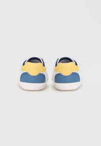 Λευκά αθλητικά παπούτσια με μπλε και κίτρινες λεπτομέρειες, με λεία δερμάτινη υφή, στρογγυλεμένο σχήμα και λευκά κορδόνια, σε προβολή από πίσω.
