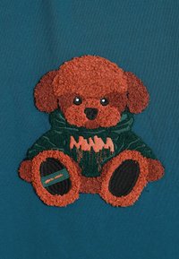 Bordado de un oso de peluche marrón sobre tela verde azulado, vestido con una sudadera verde con acentos naranjas. La textura es esponjosa, con rasgos faciales y patas prominentes.