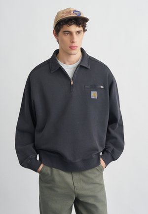 Joven con sudadera gris oscuro con cremallera hasta la mitad, bolsillo en el pecho y logo, pantalones verde oliva y gorra beige con parches.