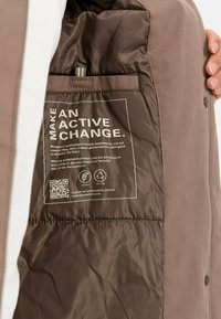 El interior de la chaqueta acolchada marrón presenta una etiqueta de cuidado con texto, un código QR y iconos de sostenibilidad. Textura suave con costuras visibles.