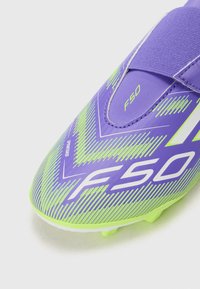 Scarpa da calcio viola con motivo zigzag verde e dettagli bianchi. Presenta una fascia sicura e una superficie testurizzata per una migliore aderenza. Modello F50.