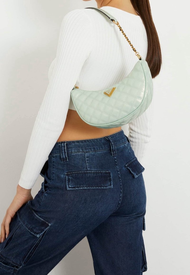Femme portant un top blanc court côtelé à manches longues et un jean cargo bleu foncé, portant un sac bandoulière matelassé vert clair en forme de croissant.