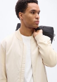 Νεαρός άνδρας με κρεμ μπουφάν τύπου bomber και λευκό πουκάμισο κρατά μια μαύρη τσάντα στον ώμο του, κοιτάζοντας δεξιά σε λευκό φόντο.
