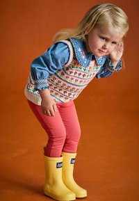Stickad väst med blommönster över en denimskjorta, parat med rosa leggings och gula gummistövlar. En orange bakgrund fullbordar scenen.