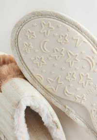 Zapatilla de tela beige con forro de piel sintética. La suela presenta un patrón en relieve de estrellas y lunas. Textura suave y diseño fácil de poner.