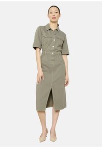 Abito in denim khaki con maniche corte, bottone frontale, due tasche sul petto, vita con cintura e spacco frontale, abbinato a tacchi beige.