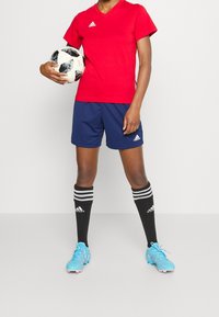 Röd v-ringad fotbollströja, marinblå shorts, svarta strumpor med vita ränder och blå fotbollsskor. Spelaren håller i en svart- och vitmönstrad fotboll.