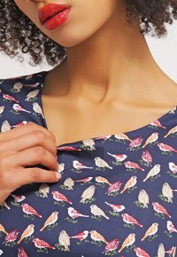 Kobieta z kręconymi włosami, ubrana w granatową bluzkę w kolorowe ptaki, trzyma materiał bluzki blisko kołnierza jedną ręką.