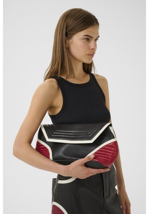 Vrouw met steil bruin haar draagt een zwart mouwloos topje en houdt een grote zwarte leren clutch met rode en witte accenten vast.