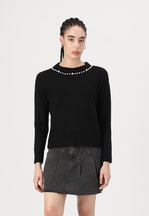 Pull en tricot noir à col rond avec une rangée décorative de perles transparentes et de petites touches florales. Associé à une jupe en denim.