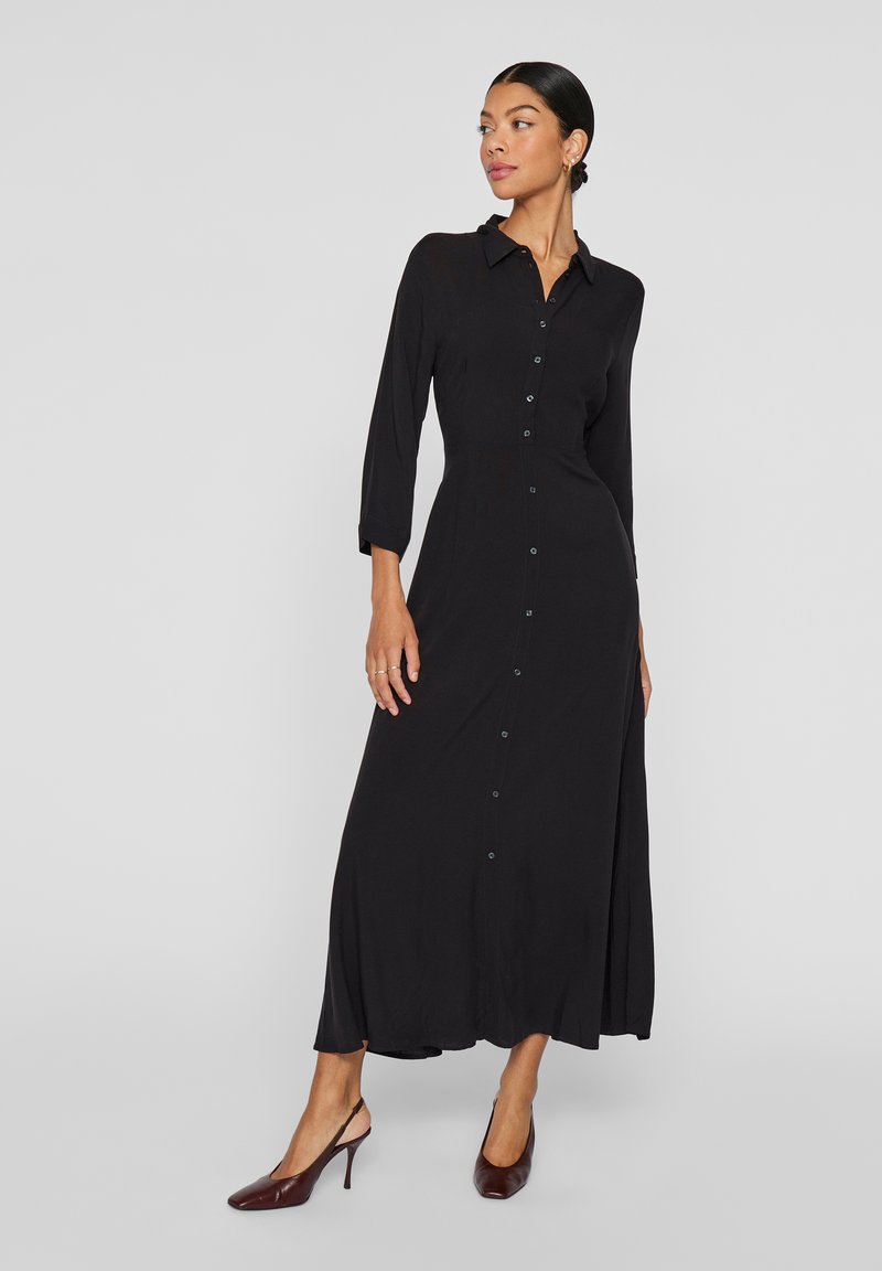 Black, long-sleeve shirt dress with buttons down the front, a pointed collar, and a flowing skirt. Paired with dark brown slingback heels.