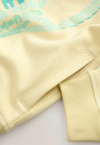 Gelbes Sweatshirt mit weicher Textur, ausgestattet mit türkisfarbigem grafischem Text und gerippten Bündchen. Enthält ein Seitennähentag für die Markenkennzeichnung.